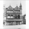 Image: a three storey commercial building on a street corner. The building has arched windows on its upper floors and large floor to ceiling display windows on the ground floor, a verandah, twin gable roofs and a corner tower.