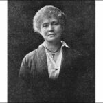 Image: Black and white photograph of woman wearing a dark jacket over a white blouse with a cross around her neck. Her light coloured hair is either pinned back or cut short.