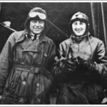 Image: A man and woman wearing First World War-era pilot’s clothing and headgear stand in front of a biplane