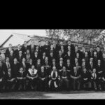 Image: A group of men in mid-20th century suits and academic gowns pose for a photograph outside a large, singles-storey stone building