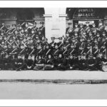 Image: Members of a large band, dressed in dark uniforms with white sashes, pose in lines with their instruments outside of a building with arched doorways.