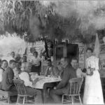 Image: A group of men and women are gathered at a table outside a timber structure. An ‘awning’ made of plant material is positioned above the table