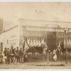 Image: a group of men, women and children in 1860s attire stand outside a single storey building which is operating as a butchers. A range of whole animal carcases are hung outside the store while other cuts of meat can be seen in a window display.