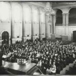 Image: A woman in cap and gown stands on a stage in front of a large audience of women dressed in late Victorian-era attire