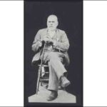 Image: A full-length photographic portrait of an elderly, bearded man sitting in a chair. The man is wearing a suit and holding a cane in his hands