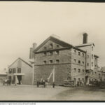 Image: A rectangular five-storey building with peaked roof sits on the edge of a dirt road. People and two horse-drawn carts stand in front of and around the building