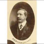 Image: A man sporting a waxed handlebar moustache and three-piece suit poses for an official photographic portrait