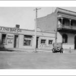 Image: A 1930s-era automobile is parked on a street in front of two buildings. One building is two storeys, made of brick, and has a second-floor verandah