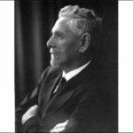 Image: A photographic head-and-shoulders portrait of a white-haired man with goatee. He is wearing a suit and is standing with his arms crossed