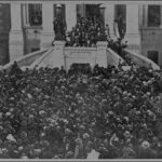 Image: a large crowd swarms the pavement in front of a twin curved staircase which leads up to a stone building with columns.