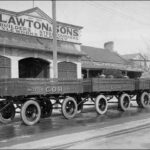 Image: three four wheeled trailers are parked in a row outside of a single storey building with arched windows and open entranceway. A parapet sign on the building reads: "J.A. Lawton & Sons. Coach builders, Steelfounders, Axelworks, Kilkenny"