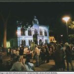 Image: a crowd gathers in front of a gothic style building which is lit up by a projection and glows purple and green