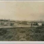 Image: A group of single-storey prefabricated buildings are situated on a remote piece of scrubby land. Low hills are visible in the distant background