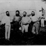 Image: four bearded men stand in front of a covered wagon