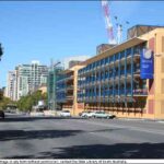 Image: a view along a main city street which is dominated by colourful four storey buildings of red and yellow with blue balconies.