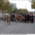 Image: three young Indigenous men in traditional dress carrying a large receptacle with fire lead a procession of people marching behind a sign featuring an Aboriginal flag and boomerang.