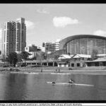 Image: rowers travel down a wide river with grassy banks in front of a 24 storey skyscraper and a large convention centre with a multi-storey curved glass wall.