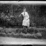 Image: A woman holding a book sits in a garden with several flowering and other plants