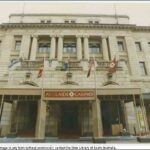 Image: a variety of flags fly above the front awning of a large, four storey stone building with arched doorways and an inset double floor balcony on its middle floors which is decorated with paired columns.