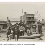 Image: A group of children sit on a merry-go-round (or joy wheel) in a park. A street and row of buildings are visible in the background