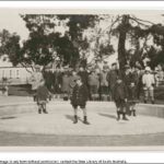 Image: A group of children pose for a photograph in an empty wading pool in a park. A street and row of buildings are visible in the background