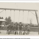 Image: A group of children pose for a photograph under a swing in a park. A building and trees are visible in the background