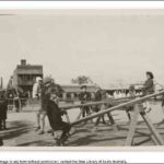 Image: A group of children play on see-saws in a park. A street and row of buildings are visible in the background