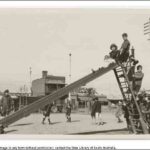 Image: A small group of children play on a slide in a park. A street and row of buildings are visible in the background