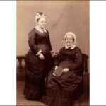 Image: sepia photograph of two women, one seated, one standing, wearing black dresses with white lace collars and white bonnets. The seated woman, the elder of the two, holds a book on her lap.