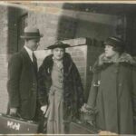 Image: A theatrical still of a young woman looking with disapproval at a young couple. The group is posed together outside the front porch of a smart house of the Art Deco period