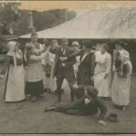 Image: A theatrical still of a group of people looking on with concern as two young women attempt to restrain a man from hitting another man who has fallen to the ground in a fight. In the background is a country homestead