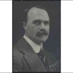 Image: A photographic head-and-shoulders portrait of a moustachioed young man in a suit and tie