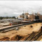 Image: a 1980s era diesel train is stopped at a platform amidst a large construction site. Behind it cranes work on a new building.