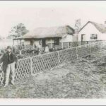 Image: A man in a bowler hat leans against a fence surrounding a garden. A small cottage connected to the garden is in the background