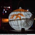 Image: A large white and orange paper lantern in the shape of a teapot glows in the dark as it is carried by a group of people in front of a stage lighting rig illuminated in pink and a banner reading moon lantern festival.