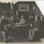 Image: photograph of seven men sitting around a table with an eighth standing behind. The photograph is torn in the top right corner.