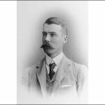 Image: A photographic head-and-shoulders portrait of a young Caucasian man wearing a light-coloured, late-Victorian era suit and sporting a very large handlebar moustache