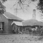 Image: man stands with dog in front of a brick house with a thatched roof. Beside him sits a woman while another man drives a horse drawn buggy.