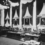 Image: a group of men sit at ornate wooden desks in a room lined with white columns and decorated with portraits, dark coloured curtains and swathes of dark fabric hanging from the ceiling.