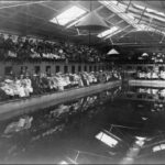 Image: A crowd of men and women in late Victorian attire sit on benches around a large swimming pool