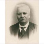 Image: A photographic head-and-shoulders portrait of a middle-aged man wearing a suit and wire-rimmed spectacles