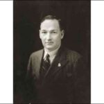 Image: A photographic head-and-shoulders portrait of a young, clean-shaven man in a dark, pinstriped suit
