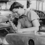 Image: A woman uses a screwdriver to assemble a large metal container. Several similar containers are arrayed in the background