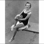 Image: A young woman in bathing attire sits on a diving board above a large swimming pool