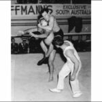 Image: A wrestler jumps on the back of his competitor while another man acting as referee looks on