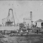 Image: A small group of men stands in front of a handful of buildings interspersed with industrial machinery