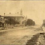 Image: a wide dirt road, torn up from traffic and littered with horse manure. A variety of small buildings line it, spaced widely apart.