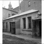 Image: A low, rectangular stone and brick building surrounded on three sides by much taller brick and stone buildings. The back door of the building features a wooden latticework awning