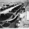 Image: Black and white photograph of a tree lined boulevard, as viewed from the roof of a building. The buildings lining one side of the road are grand stone constructions and are surrounded by formal gardens.