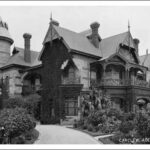 Image: A large, two-storey stone mansion with a cylindrical turret comprising one corner of the structure